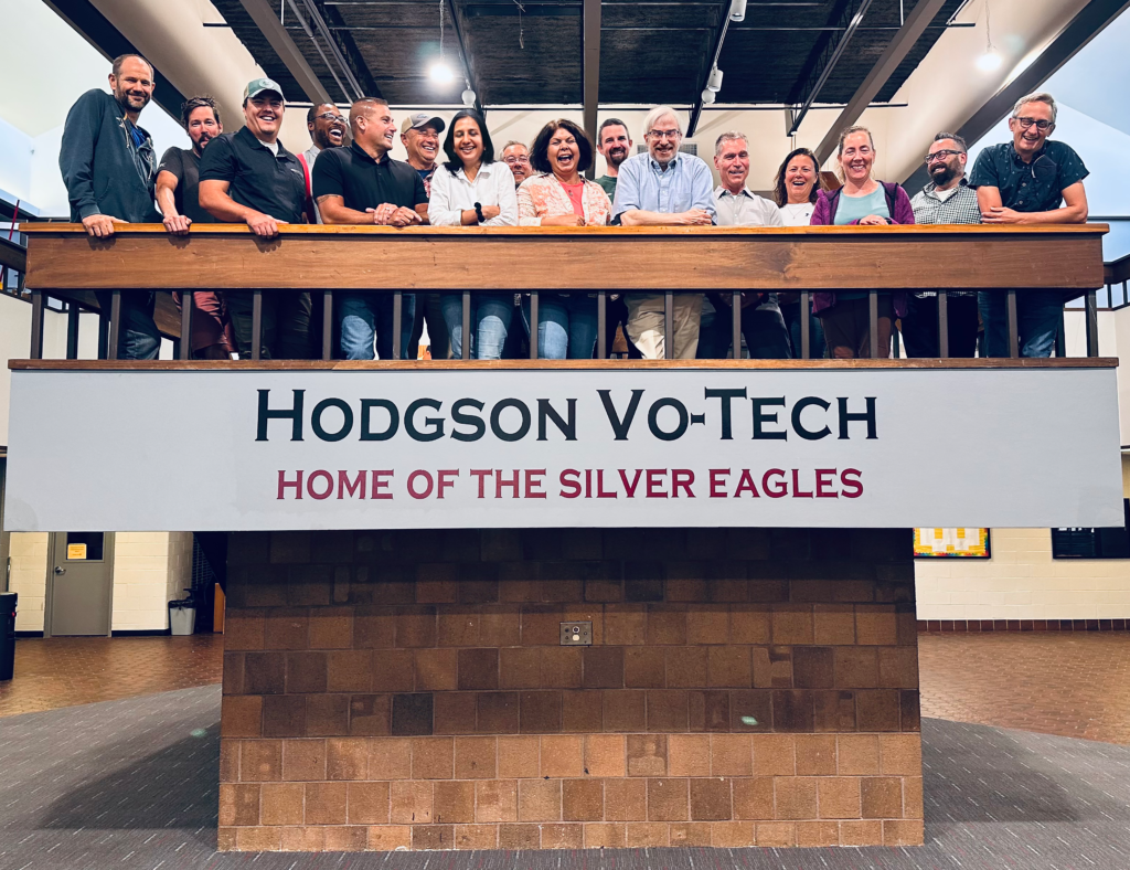 A group standing on a platform with a sign saying "Hodgson Vo-Tech, Home of the Silver Eagles"